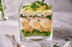 a salad with hard boiled eggs, noodles and greens in a square glass container next to a spoon