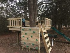 a child standing on top of a wooden play structure next to a tree with a slide