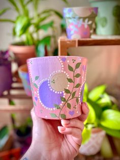 a hand holding a pink cup with flowers painted on it and green plants in the background