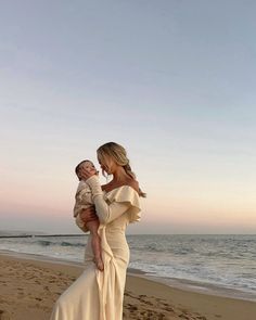 a woman holding a baby standing on top of a sandy beach next to the ocean