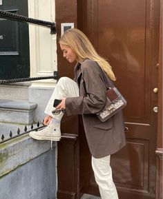 a woman in white pants and brown jacket leaning against a door with her cell phone