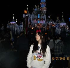 a woman standing in front of a castle with lights on it's sides and people walking around