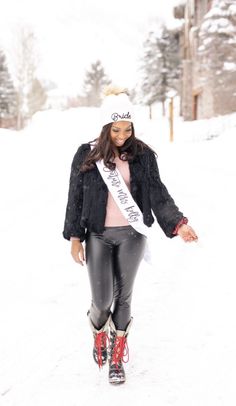 a woman is walking in the snow wearing black leather pants and a white hat with a sash around her neck