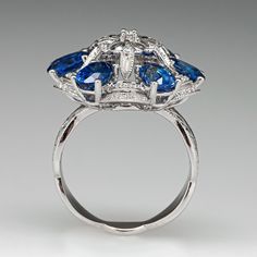 This exquisite cluster style ring is centered with a pierced floral cluster setting accented with seven (7), bead/prong set, round brilliant cut diamonds and twenty-eight (28), bead set, round brilliant/single cut diamonds, bordered with six (6), prong set, oval mixed cut natural sapphires and forty-two (42), bead set, round brilliant cut diamonds. The diamonds are bordered with milgrain edging. Engraved details accent the shoulders of the shank. The ring measures 21.3mm at the top, rises 10.2mm above the finger, tapering to 3.8mm wide and 1.0mm thick at the base of the shank. This ring is currently a size 6.75. Luxury Cluster Diamond Ring With Gemstone, Luxury Sapphire Cluster Ring With Brilliant Cut, Luxury Diamond Cluster Ring With Gemstones, Luxury Cluster Ring With Round Cut Gemstone, Luxury Cluster Sapphire Ring With Center Stone, Luxury Blue Topaz Rings With Rose Cut Diamonds, Luxury Blue Diamond Ring With Accents, Luxury Cluster Ring With Sapphire In Round Cut, Luxury Sapphire Cluster Ring With Gemstones