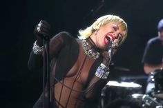 lady in sheer bodysuit singing into microphone with band on stage behind her at concert