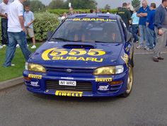 a blue subaru racing car with yellow lettering on it's hood is parked in front of a group of people