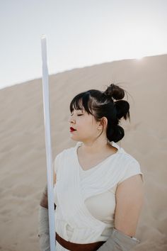 a woman in white dress holding a pole and standing in the sand with her eyes closed