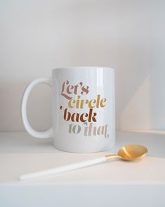 a white coffee mug with gold lettering on it next to a spoon that says let's circle back no time