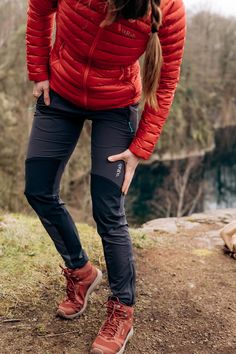 a woman in red jacket and black pants standing on top of a hill with her back to the camera