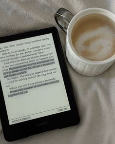 a cup of coffee sitting next to a kindle on a white blanket with a book in front of it