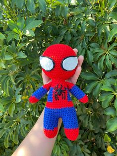 a hand holding up a crocheted spiderman doll in front of some bushes