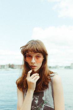 a woman wearing glasses standing next to the water with her hand on her chin and looking off into the distance