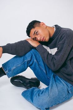 a young man sitting on the ground with his hand under his chin, wearing jeans and a sweater