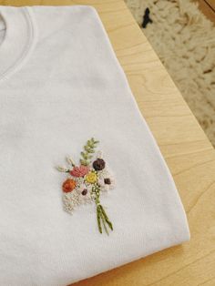 a white t - shirt with flowers embroidered on the front and back, sitting on a wooden table