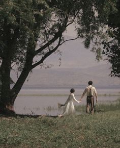 a man and woman holding hands while walking by the water with trees in front of them