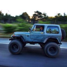 a blue jeep driving down the road with trees in the backgrouund and one person sitting in the driver's seat