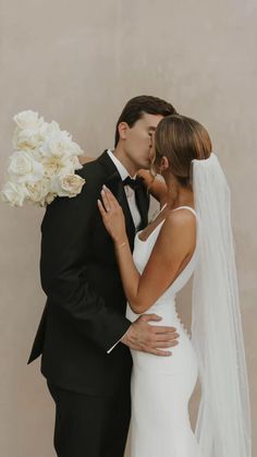 a bride and groom kissing each other in front of a beige wall with white flowers