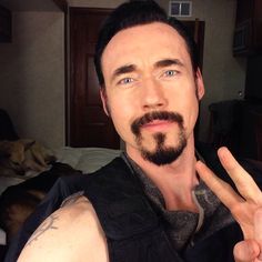 a man with a goatee making the peace sign while sitting on a bed in his room