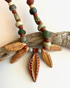 "The wonderfully textured stoneware leaves and pods in this necklace were hand formed and fired by another Etsy artisan.  I added color, a soft golden brown for the four smaller dangles, 35-49mm, and a deeper yellow-brown with touches of coral for the central pod, 52mm. They are strung with orange-red, green, and tan beads in a variety of shapes and materials, including old glass, ceramic, stone, and bone. Most of the beads are matte, earthy, slightly rustic, with muted colors. Ceramic beads in Nature-inspired Necklace With Large Beads For Gifts, Unique Red Necklaces With Dangling Beads, Unique Red Necklace With Dangling Beads, Red Natural Stones Nature-inspired Jewelry, Handmade Red Nature-inspired Necklace, Nature-inspired Jewelry With Dangling Beads For Gifts, Earthy Red Handmade Jewelry, Earthy Red Jewelry With Natural Stones, Earthy Red Jewelry For Gifts