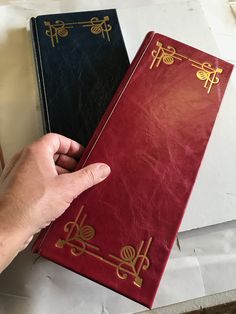 a hand is pointing at two red and black books with gold designs on the covers