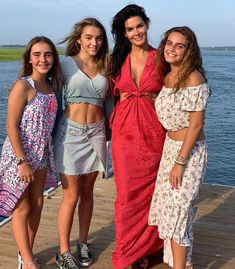 three women standing on a dock next to the water with one woman wearing a red dress