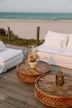 two coffee tables sitting on top of a wooden deck next to the ocean with white couches and pillows