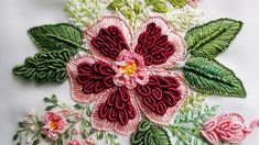 an embroidered flower with green leaves and pink flowers on white fabric, in the middle of embroidery work