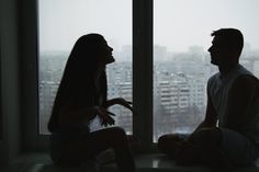 a man and woman sitting next to each other in front of a window looking out at the city