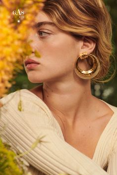 a woman wearing large gold hoop earrings in front of yellow flowers with the caption caas