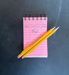 a pink notepad with two pencils next to it on a blackboard background