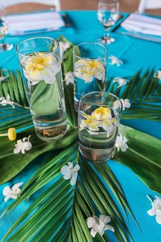 three glasses filled with water on top of a blue table cloth covered in white and yellow flowers