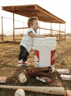 a little boy standing next to a bucket full of baseballs and ball bats on the ground