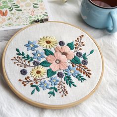 a cup of coffee next to a flower embroidered on a white table cloth with a blue mug in the background