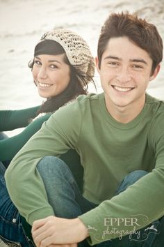 a young man and woman are sitting on the beach smiling at the camera with their arms around each other