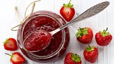 strawberry jam in a glass jar with spoons and strawberries next to it on a white wooden table