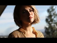 a woman with blonde hair wearing a gold jacket and looking off into the distance while standing in front of a tree