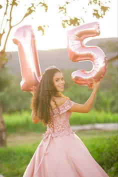 a woman in a pink dress holding up balloons that spell the number five, and wearing a tiara