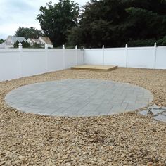 a circular stone patio surrounded by gravel and white fence with trees in the back ground