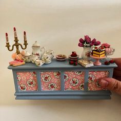 a hand holding a miniature tea set on top of a blue table with pink and white flowers