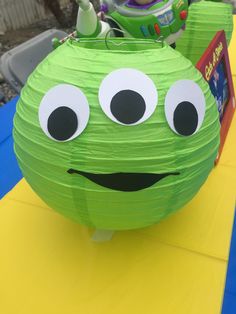 a green paper lantern with googly eyes on it's face and mouth, sitting on top of a yellow table