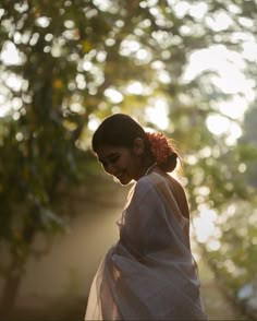 a woman wearing a white sari standing in front of some trees and sunlight shining through the leaves