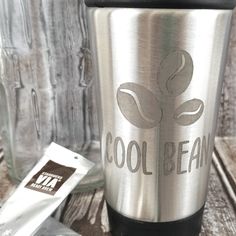 a stainless steel coffee cup sitting on top of a wooden table next to a bag of beans