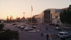 the sun is setting over a parking lot with cars parked in front of it and people walking around