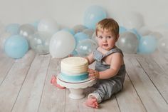 a little boy sitting on the floor with a cake in front of him and balloons behind him