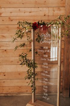 a wedding ceremony sign with greenery and flowers on it, sitting in front of a wooden wall