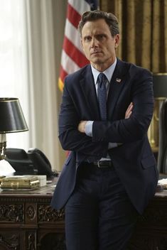 a man in a suit and tie standing next to a desk with an american flag on it