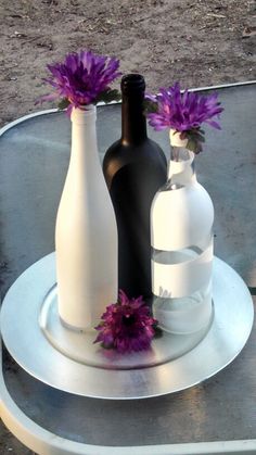 three white vases with purple flowers in them sitting on a metal tray near an empty bottle