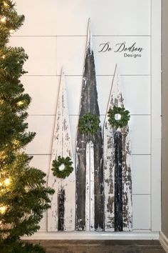 a christmas tree is in front of a wall with three wooden trees on it and wreaths hanging from the top