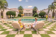 two women sitting on lawn chairs in front of a swimming pool with palm trees around it