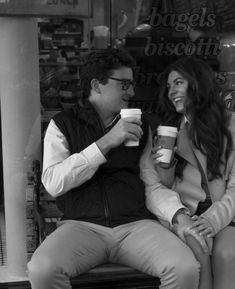 a man and woman sitting on a bench holding coffee cups in front of a store window